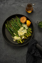 Fried green asparagus with lemon and parmesan in a pan.  Black marble table with olive oil and pesto. Top view.