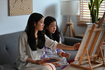 A portrait of happy Asian girls sitting in a living room holding a paint brush, color palette and working on a canvas together, for art, education, home and family concept.
