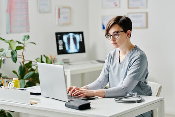 Young serious female doctor in medical scrubs typing on laptop keyboard while consulting online...