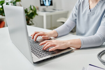 Hands of young female physician or other specialist in blue uniform typing on laptop keyboard while sitting by workplace