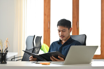 A portrait of a young Asian man sitting in the office holding and looking at the report, data and documents, for business and  technology concept.