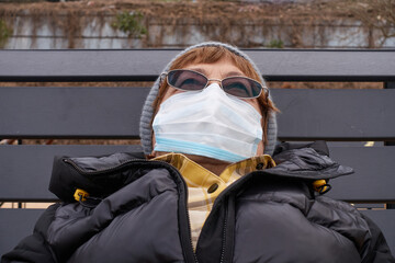 Grandmother in glasses and a protective mask reclining is resting on a bench in a winter park. Selective focus.