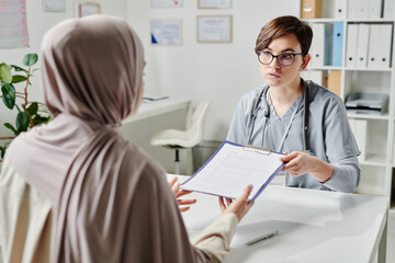 Young serious medical staff passing document in clipboard to Muslim female patient in hijab during...