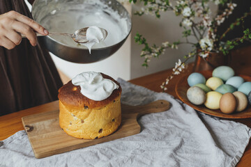 Woman decorating homemade easter bread with sugar glaze on rustic table with natural dyed eggs and spring blossom in room. Happy Easter! Woman baking stylish easter cake
