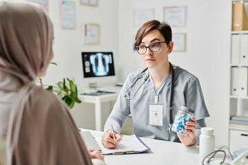 Confident doctor in medical scrubs showing bottle of pills to Muslim female patient in hijab while prescribing her effective medicament