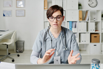 Confident doctor in medical scrubs communicating to online patients while sitting by desk in office...