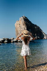 A beautiful young woman in a hat and a light dress with her back is walking along the ocean shore against the background of huge rocks on a sunny day. Tourism and vacation travel.