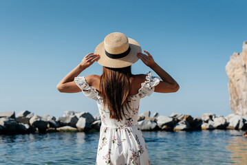 A beautiful young woman in a hat and a light dress with her back is walking along the ocean shore against the background of huge rocks on a sunny day. Tourism and vacation travel.