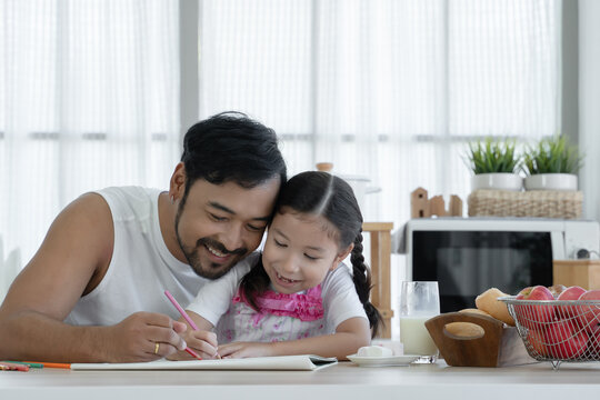Happy Asian young father with beard and little cute daughter enjoy drawing with colored pencil together at home with glass of milk, marshmallows, breads and red apples on kitchen table