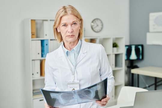 Serious mature female doctor with x-ray image of patient lungs looking at camera while standing against shelves and computer in medical office