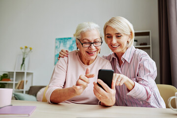 Portrait of smiling young woman helping senior mother using smartphone in cozy home interior