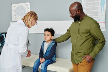 Happy young African American man standing by his cute little son and looking at him in clinics...
