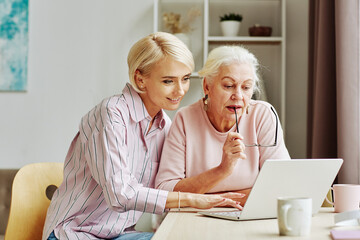 Portrait of blonde young woman teaching senior mother using laptop and internet in cozy home...