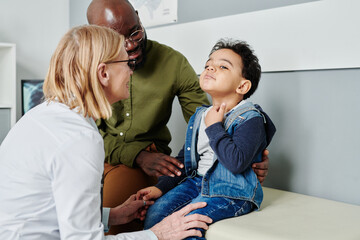 Sick little boy complaining on sore throat to mature female peditrician while sitting on bench next to his father during visit to doctor