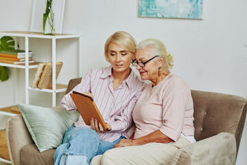 Portrait of blonde young woman helping senior mother using digital tablet and online services while sitting on couch at home together