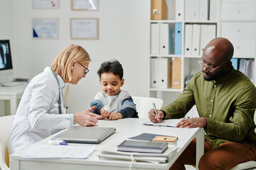 Young African American man filling in medical form while sitting by desk in front of his little son and doctor using pulse oximeter