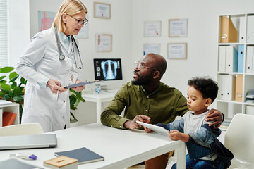 Mature female doctor in whitecoat standing by workplace and consulting young African American man with little son in clinics