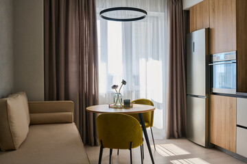 Spacious kitchen in modern apartment with table and chairs standing in the center surrounded by beige sofa, refligerator and oven