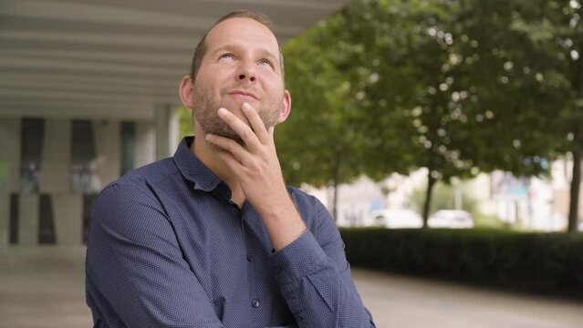 A Caucasian man thinks about something by an office building in an urban area - closeup - a street in the blurry background