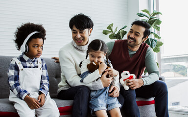 Banner with copy space couple male gay taking care with love to adopted children who are little diverse Caucasian and African girl and boy, sitting on sofa in living room at home. LGBT, kids Concept.