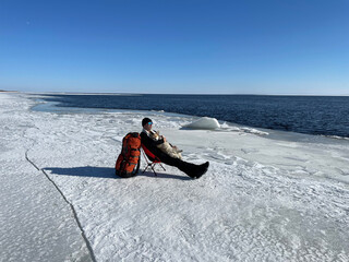 Fototapeta na wymiar Hiker with cute dog corgi on hands sitting in tourist chair on ice of Lake Ladoga shore and enjoying the view of North Karelia in early spring