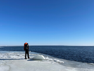 Hiker with backpack standing on ice of Lake Ladoga shore and enjoying the view of North Karelia in early spring on sunny day