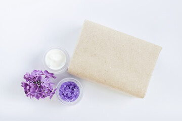 Soap in craft paper, sugar scrub, cream with lilac flowers on white background. Mockup and copy space. Spa treatments for home care. Home rejuvenation and moisturizing