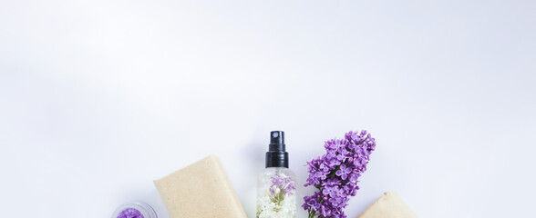 Spa treatments for home care. Home rejuvenation and moisturizing. Banner with soap in craft paper, aroma oil, sugar scrub, cream with lilac flowers on white background. Mockup and copy space.