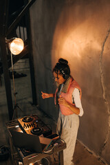 Stylish girl in casual clothes dances and plays DJ set at the party, uses the controller and laptop.