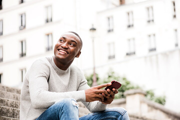 smiling African american man sitting outside in city holding cellphone