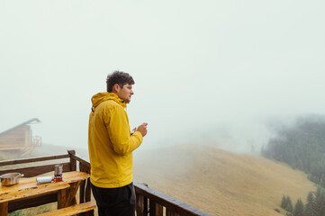 A man in a yellow jacket spends time at a dacha in the mountains, standing on the terrace with a...