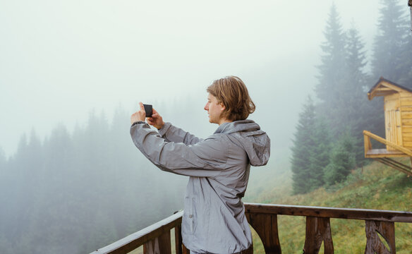 Male tourist stands on the terrace of a wooden house in the mountains on a foggy morning and takes photos of beautiful landscapes on a smartphone camera.