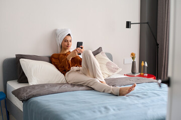 Young restful female in home wear sitting on double bed and texting in mobile phone after morning shower in hotel room or bedroom