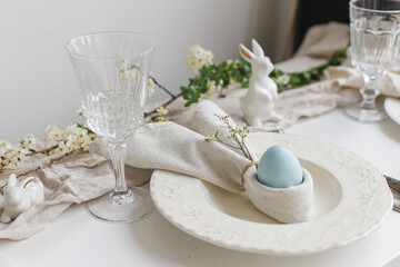 Easter egg in bunny napkin on plate with cutlery, bunny, spring flowers on rustic white table. Stylish elegant Easter brunch table setting. Modern table arrangement and eco decoration