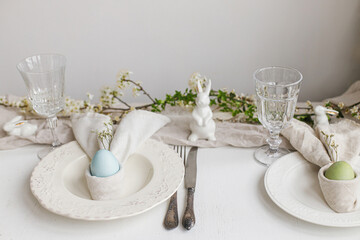Easter egg in bunny napkin on plate with cutlery, bunny, spring flowers on rustic white table. Stylish elegant Easter brunch table setting. Modern table arrangement and eco decoration