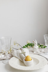 Happy Easter! Stylish elegant Easter brunch table setting. Easter egg in bunny napkin on plate with cutlery, bunny, spring flowers on rustic white table. Modern eco table arrangement