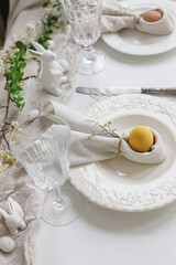 Happy Easter! Stylish elegant Easter brunch table setting. Easter egg in bunny napkin on plate with cutlery, bunny, flowers on rustic white table. Modern eco table arrangement. Top view