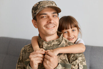 Indoor shot of positive young adult man soldier wearing camouflage uniform and cap, posing with his...