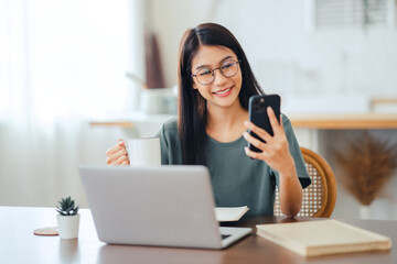 Happy asian woman works at home and uses a smart phone and a notebook laptop computer on table. Smiling young female wearing eyeglasses messaging with smartphone. - obrazy, fototapety, plakaty