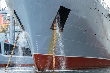 Close-up of the bow of the gray hull of a warship. Rusty chains hold anchors. Water is pouring out...