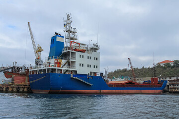 Fototapeta premium An old blue ship is in port for loading. In the background are booms of cranes and a cloudy sky.
