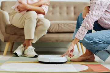 Cropped shot of young woman pushing button on robot vacuum cleaner while showing smart home technology to senior relative, copy space