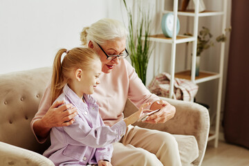 Side view portrait of smiling little girl showing mobile phone to grandmother and teaching her...