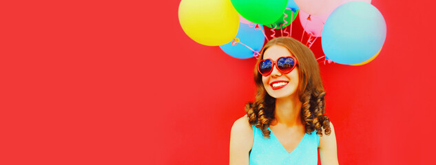Portrait of happy smiling young woman with colorful balloons wearing heart shaped sunglasses on red background