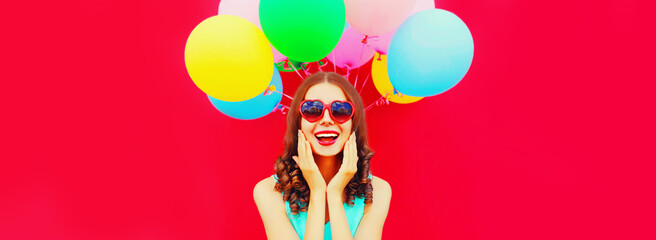 Portrait of happy smiling young woman with colorful balloons wearing heart shaped sunglasses on red background