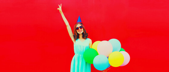 Obraz na płótnie Canvas Happy cheerful smiling young woman in birthday hat with colorful balloons on pink background, blank copy space for advertising text