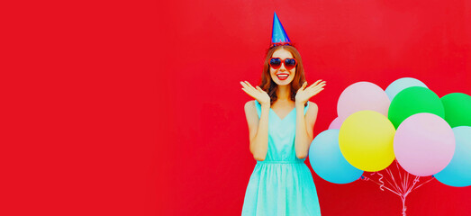 Obraz na płótnie Canvas Portrait of happy smiling young woman with colorful balloons wearing birthday hat on red background, blank copy space for advertising text
