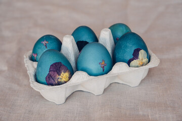 Naturally dyed cyan blue pressed flower Easter eggs in egg box on neutral beige tablecloth background. Handmade craft natural decoration.
