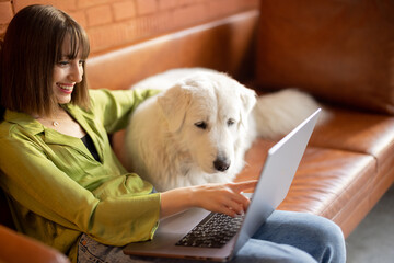 Young woman works on laptop computer while sitting with her cute white dog on a couch at home. Remote work from home and friendship with pets