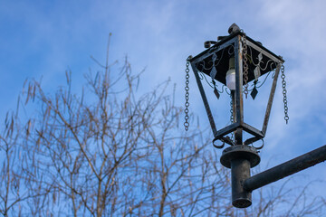 Fototapeta na wymiar Black iron street lamp against the background of trees and blue sky. Forged details of architecture. Vintage garden lantern. Metal objects in art.
